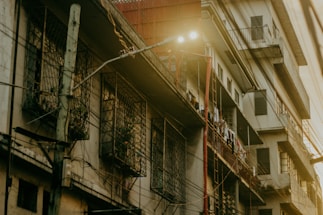 An urban residential building with a slightly worn exterior shows balconies and window grilles. Clothes are hanging to dry on the railings. Electric wires run across the facade, and a streetlamp emits light nearby, casting an atmospheric glow.
