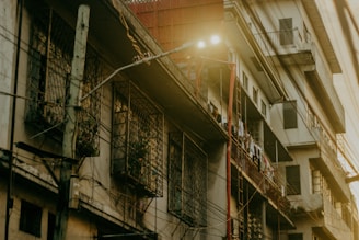 An urban residential building with a slightly worn exterior shows balconies and window grilles. Clothes are hanging to dry on the railings. Electric wires run across the facade, and a streetlamp emits light nearby, casting an atmospheric glow.