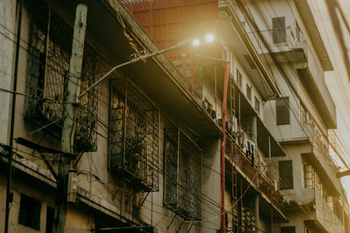 An urban residential building with a slightly worn exterior shows balconies and window grilles. Clothes are hanging to dry on the railings. Electric wires run across the facade, and a streetlamp emits light nearby, casting an atmospheric glow.