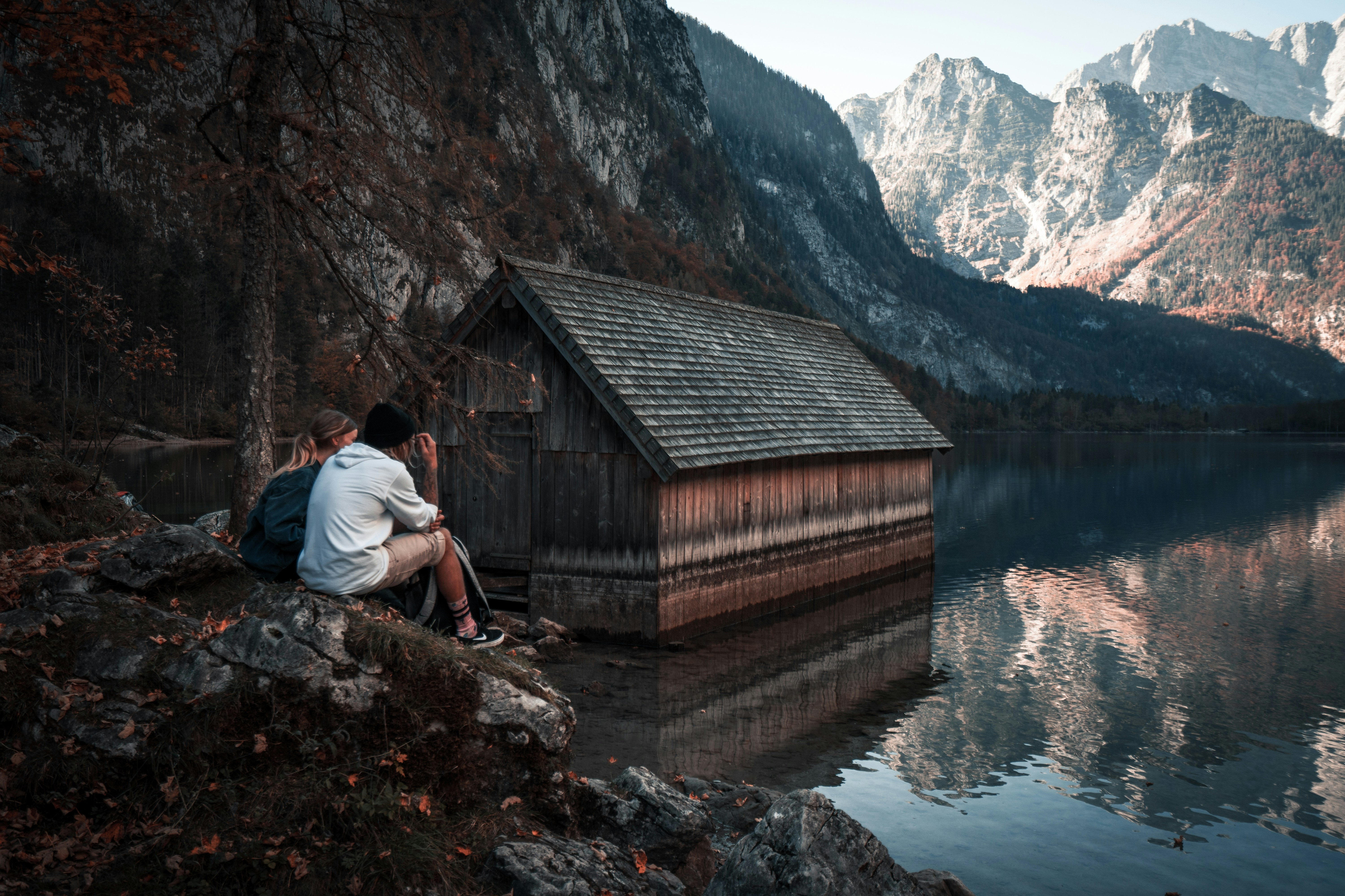 a couple of people sitting on a rock ledge looking at a cabin in the mountains, Couple sitting at the lake 