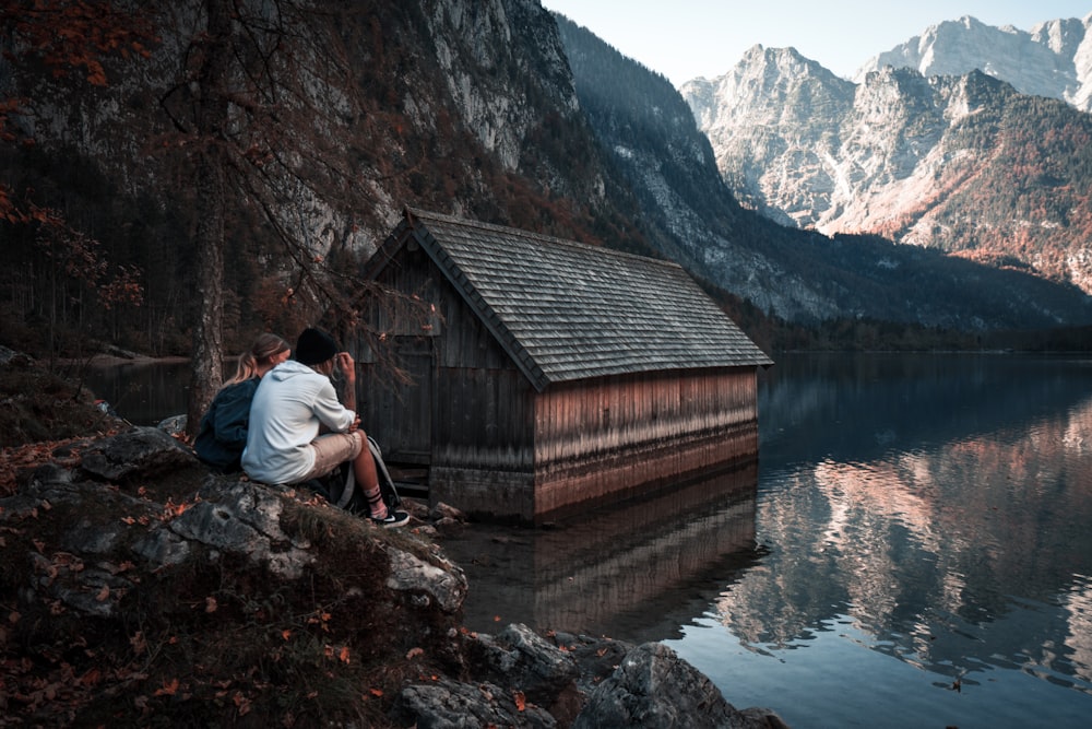 a couple of people sitting on a rock ledge looking at a cabin in the mountains