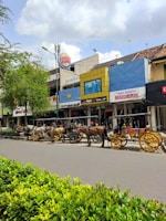 A street scene with several horse-drawn carriages lined up along a sidewalk in front of various shops. The carriages feature colorful decorations and yellow wheels. The shops have signs with names like Burger King and Toko Sepatu Modern, and trees and bushes are visible along the street. The sky is cloudy but the atmosphere looks bright.