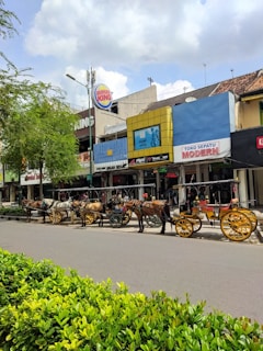 A street scene with several horse-drawn carriages lined up along a sidewalk in front of various shops. The carriages feature colorful decorations and yellow wheels. The shops have signs with names like Burger King and Toko Sepatu Modern, and trees and bushes are visible along the street. The sky is cloudy but the atmosphere looks bright.