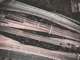 An aerial view of a highway interchange with multiple lanes of traffic, including several cars moving in different directions. The roads are elevated and lined with greenery, and there are visible markings on the road indicating lanes and directions.