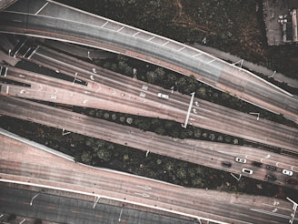 An aerial view of a highway interchange with multiple lanes of traffic, including several cars moving in different directions. The roads are elevated and lined with greenery, and there are visible markings on the road indicating lanes and directions.