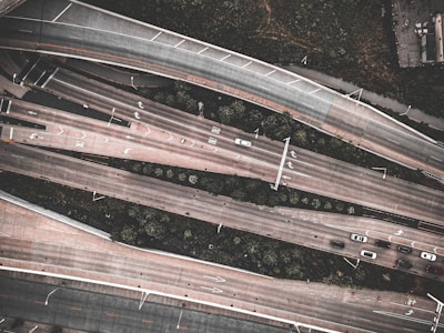 An aerial view of a highway interchange with multiple lanes of traffic, including several cars moving in different directions. The roads are elevated and lined with greenery, and there are visible markings on the road indicating lanes and directions.