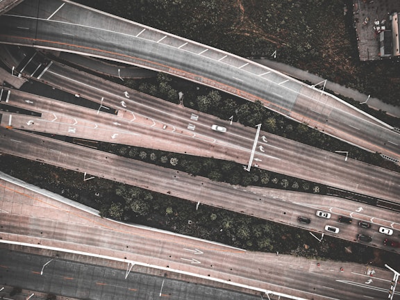 An aerial view of a highway interchange with multiple lanes of traffic, including several cars moving in different directions. The roads are elevated and lined with greenery, and there are visible markings on the road indicating lanes and directions.