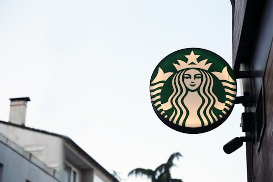 A round green and white sign with an image of a mermaid or siren, typically associated with a popular coffee chain, mounted on the side of a building. The background features parts of a building with windows and trees visible in the distance.