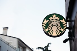 A round green and white sign with an image of a mermaid or siren, typically associated with a popular coffee chain, mounted on the side of a building. The background features parts of a building with windows and trees visible in the distance.