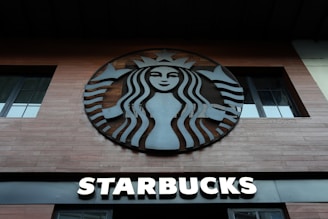 A wooden-planked storefront features a large, circular logo with a stylized siren in black and white. Below the logo, bold letters spell out 'Starbucks' in illuminated white typography.