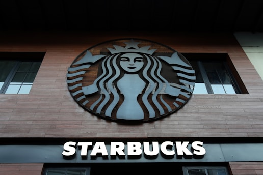 A wooden-planked storefront features a large, circular logo with a stylized siren in black and white. Below the logo, bold letters spell out 'Starbucks' in illuminated white typography.
