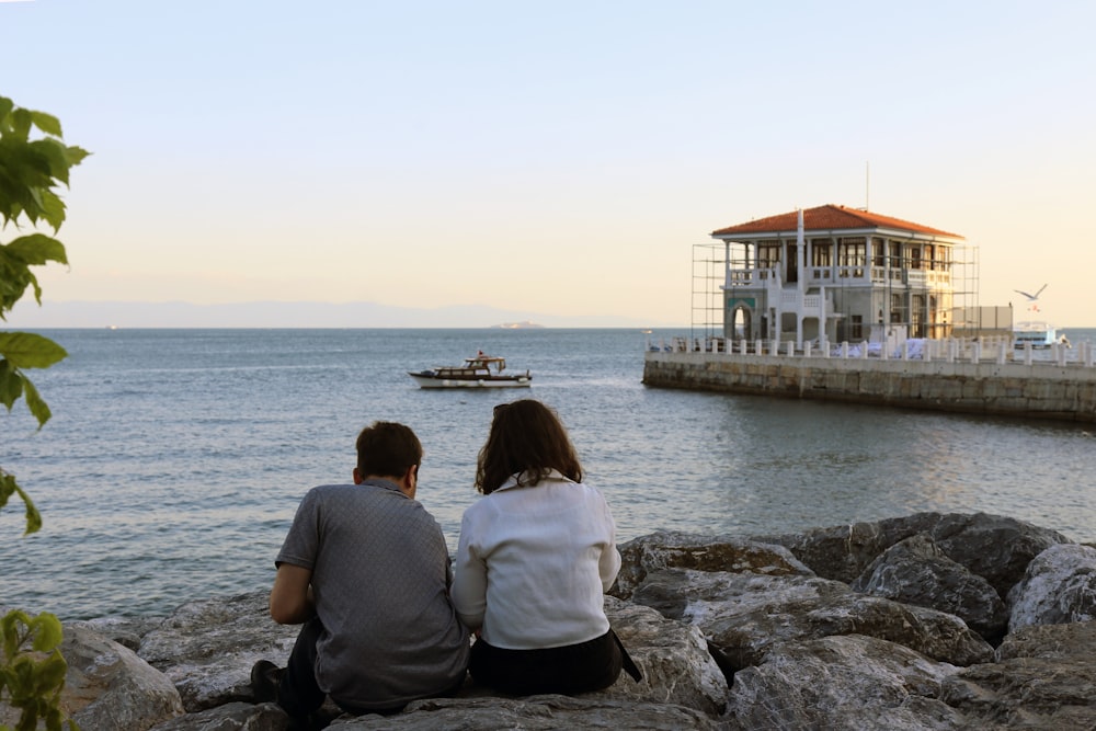 a couple sitting on rocks looking at a boat in the water