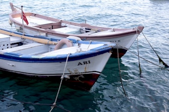 Two small wooden boats are floating on the water's surface. The water appears clean and gently rippled with shades of blue and green. One boat has a blue trim with a name painted on its side. A small red flag is hoisted at the stern of the rear boat.