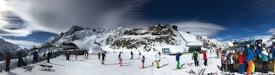 A panoramic view of a snowy mountain landscape with people skiing and snowboarding. Several skiers and snowboarders are on a snow-covered slope with ski lifts and a mountain backdrop. The sky is partly cloudy and the scene is lively with activity.