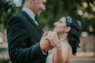 A couple is holding hands while dancing, both smiling and dressed formally. The background is blurred, featuring green tones, suggesting an outdoor setting.