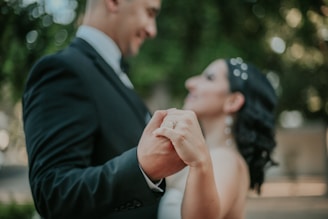 A couple is holding hands while dancing, both smiling and dressed formally. The background is blurred, featuring green tones, suggesting an outdoor setting.
