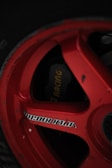 A close-up of a red racing wheel rim with visible text and a brake caliper behind it. The rim has a glossy finish and displays some wear.