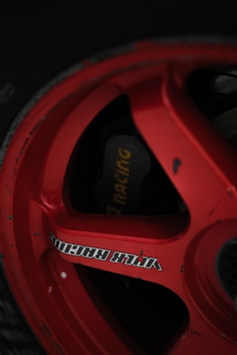 A close-up of a red racing wheel rim with visible text and a brake caliper behind it. The rim has a glossy finish and displays some wear.