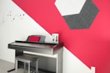 A modern music room featuring a digital keyboard with a red cushion resting against its back. The wall behind has an artistic design with red and white geometric patterns and a black hexagonal sound panel. Musical notes are depicted as a decorative element on the white section of the wall.