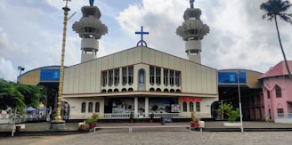 A large church with a prominent cross on the roof, flanked by two tall towers. The facade of the church is decorated with a series of arched windows and a central mural. In front of the church, there is a paved area with potted plants and a large brass lamp. The sky above is cloudy, and there are additional buildings visible on the sides.