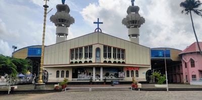 A large church with a prominent cross on the roof, flanked by two tall towers. The facade of the church is decorated with a series of arched windows and a central mural. In front of the church, there is a paved area with potted plants and a large brass lamp. The sky above is cloudy, and there are additional buildings visible on the sides.