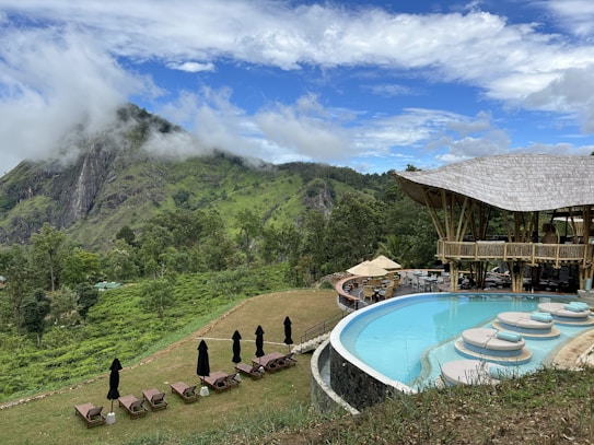 A picturesque landscape featuring a modern, open-air structure with a thatched roof and an adjacent infinity pool overlooking lush green hills and a striking rocky mountain. Several lounge chairs with closed umbrellas are arranged near the pool, and an outdoor seating area with tables and chairs is visible. The sky is partly cloudy, contributing to a serene and tranquil atmosphere.