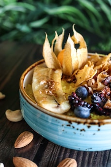 A close-up of a bowl filled with a variety of fruits and nuts, including banana slices, physalis, blueberries, and pomegranate seeds. The bowl has a rustic appearance with a blue glaze, and the background features lush greenery, adding a natural touch.