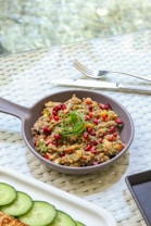A gray ceramic dish filled with a colorful mixture of cooked grains, vegetables, topped with pomegranate seeds and garnished with green herbs. The dish rests on a woven placemat next to a knife and fork. There is a white plate with sliced cucumbers partially in view.