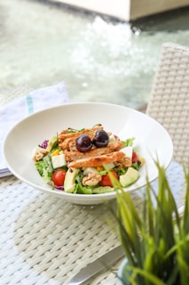 A fresh, colorful salad is served in a white bowl. The salad contains ingredients like grilled chicken strips, cherries, avocado slices, walnuts, cherry tomatoes, corn, and pieces of feta cheese on a bed of mixed greens. The setup is outdoors on a woven texture tablecloth with a blurred background featuring water and a plant.