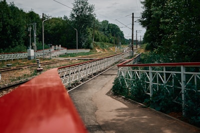 A railway track is running through a lush green area, with trees lining both sides of the track. A red and white railing runs alongside the track, leading into the distance. Overhead electric wires indicate that this is an electrified railway line. The scene is calm and slightly overcast, suggesting a tranquil rural setting.