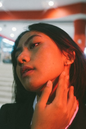 A portrait of a person with long dark hair, softly lit with warm, orange-red hues. The focus is on their thoughtful expression as they gaze upward, with one hand gently touching their neck. The blurred background features soft lights, contributing to an atmospheric ambience.