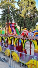A colorful amusement park ride featuring a miniature train with vibrant yellow, green, and purple colors. There are several train cars with decorative elements, such as multi-colored circular accents. In the background, large inflatable slides in bright red, green, and blue stripes rise against a backdrop of tall trees. A ticket booth is visible beside the ride.