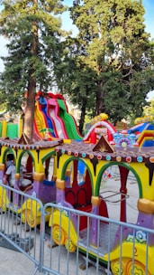A colorful amusement park ride featuring a miniature train with vibrant yellow, green, and purple colors. There are several train cars with decorative elements, such as multi-colored circular accents. In the background, large inflatable slides in bright red, green, and blue stripes rise against a backdrop of tall trees. A ticket booth is visible beside the ride.