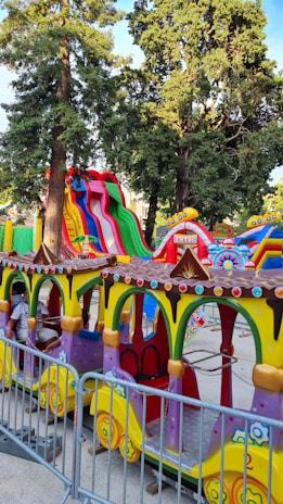 A colorful amusement park ride featuring a miniature train with vibrant yellow, green, and purple colors. There are several train cars with decorative elements, such as multi-colored circular accents. In the background, large inflatable slides in bright red, green, and blue stripes rise against a backdrop of tall trees. A ticket booth is visible beside the ride.