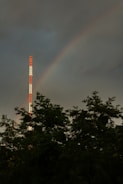 A tall radio or communication tower with alternating red and white sections is seen rising above the treetops. A faint rainbow arcs in the cloudy sky beside the tower, adding a splash of color to the otherwise dark and moody scene.