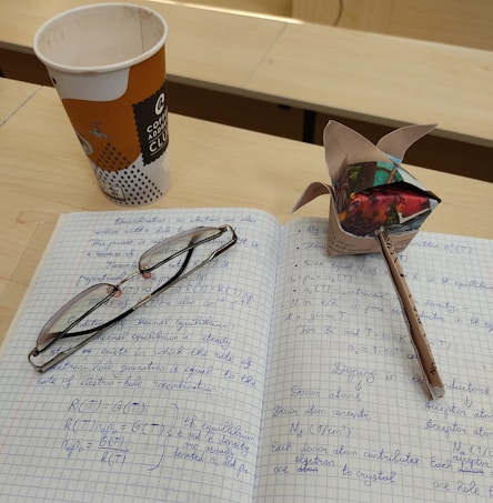 A checked notebook is open with handwritten notes covering technical content, possibly related to chemistry or physics. A pair of reading glasses rests on the notebook. Beside the notebook is a folded origami paper structure made from a magazine or colorful paper. A pencil is inserted into the origami structure. A disposable coffee cup is nearby on the table, indicating a study or classroom environment.