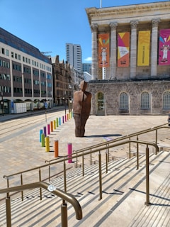 A modern art sculpture stands in front of a historic building with tall columns. Colorful poles line the area around the sculpture, and banners hang on the building promoting an event. Surrounding the scene are modern and older styled buildings.