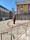 A modern art sculpture stands in front of a historic building with tall columns. Colorful poles line the area around the sculpture, and banners hang on the building promoting an event. Surrounding the scene are modern and older styled buildings.