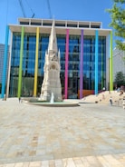 A modern building with a colorful facade featuring large vertical stripes in various hues stands behind a detailed historical-style monument with a spire. The forecourt is a spacious, tiled area where a few people are walking and sitting, enjoying the sunny weather.