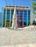 A modern building with a colorful facade featuring large vertical stripes in various hues stands behind a detailed historical-style monument with a spire. The forecourt is a spacious, tiled area where a few people are walking and sitting, enjoying the sunny weather.