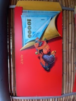 A vibrant red panel features an artistic depiction of a traditional Asian figure in colorful attire, holding a large piece of currency with the number 1000 prominently displayed. The figure is adorned with intricate designs and patterns, symbolizing prosperity or fortune. Chinese characters are written vertically on the panel.