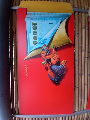 A vibrant red panel features an artistic depiction of a traditional Asian figure in colorful attire, holding a large piece of currency with the number 1000 prominently displayed. The figure is adorned with intricate designs and patterns, symbolizing prosperity or fortune. Chinese characters are written vertically on the panel.