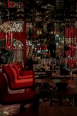 A dimly lit, elegant restaurant interior with red chairs lined up at wooden tables. Wine glasses are neatly arranged, and decorative light fixtures hang from the ceiling. The reflection patterns on glossy surfaces create an upscale ambiance.