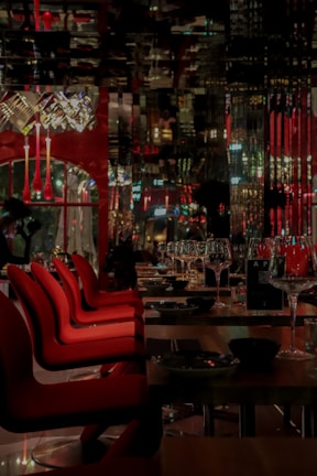 A dimly lit, elegant restaurant interior with red chairs lined up at wooden tables. Wine glasses are neatly arranged, and decorative light fixtures hang from the ceiling. The reflection patterns on glossy surfaces create an upscale ambiance.