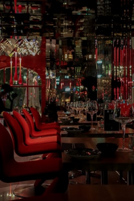 A dimly lit, elegant restaurant interior with red chairs lined up at wooden tables. Wine glasses are neatly arranged, and decorative light fixtures hang from the ceiling. The reflection patterns on glossy surfaces create an upscale ambiance.