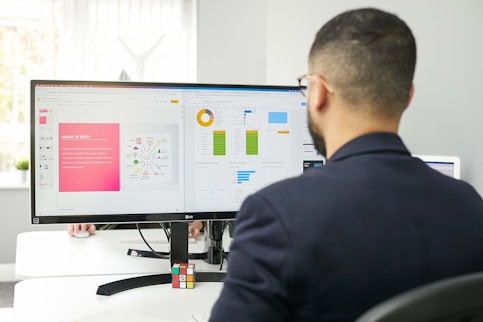 A man in a suit is working at a desk with a dual-monitor setup showing SEO presentation slides and data charts. The office environment is bright, with natural light coming from a nearby window, and a Rubik's cube is placed in front of the monitor.