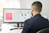 A man in a suit is working at a desk with a dual-monitor setup showing SEO presentation slides and data charts. The office environment is bright, with natural light coming from a nearby window, and a Rubik's cube is placed in front of the monitor.