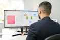 A man in a suit is working at a desk with a dual-monitor setup showing SEO presentation slides and data charts. The office environment is bright, with natural light coming from a nearby window, and a Rubik's cube is placed in front of the monitor.