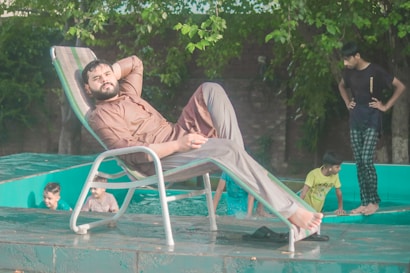 A man is relaxing on a reclining chair beside a pool, wearing comfortable brown attire. Several children and young people are playing in the pool nearby. The background is filled with lush green trees, creating a serene and casual atmosphere.
