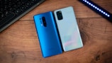 Two smartphones are placed on a wooden surface. The phone on the left is blue with a vertical camera setup, and the one on the right is a reflective, light-colored phone with a rectangular camera module. Nearby, part of a keyboard and an LED light strip are visible on the sides.